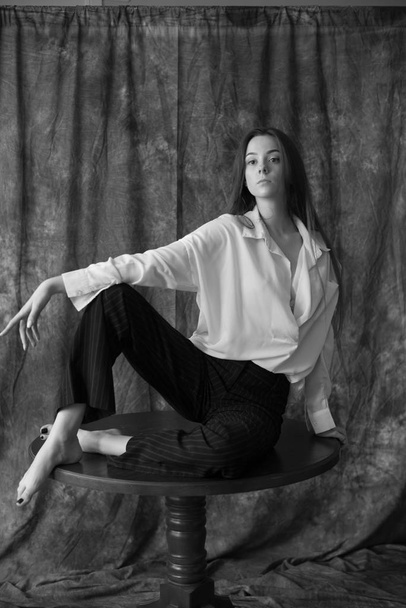 Lovely young girl model with dark long hair in a white shirt and black pants posing in the studio on a textile background. Beauty and fashion. - Fotó, kép