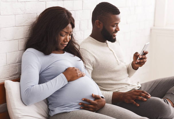 Worried pregnant woman touching tummy while indifferent husband using phone - Foto, Imagen