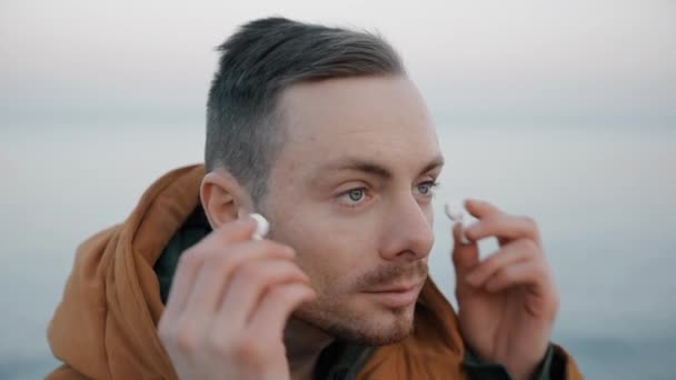 Handsome confident man puts on earphones at the sea beach - Materiaali, video