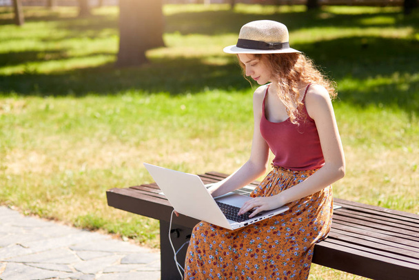 Outdoor shot of girl looking for information via laptop and wireless Internet, charges with help of solar panel built in wooden bench, looks concentrated, doing online work. Alternative energy concept - Фото, изображение