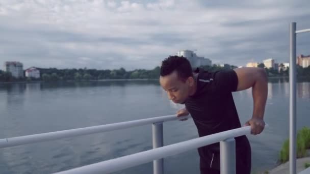 Afro man doing triceps dips on parallel bars near lake - Video, Çekim