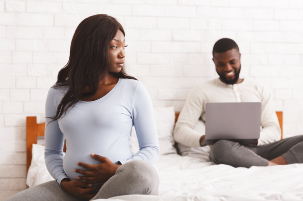 Θυμωμένη έγκυος κυρία που κάθεται στο κρεβάτι ενώ ο σύζυγος χρησιμοποιεί φορητό υπολογιστή - Φωτογραφία, εικόνα