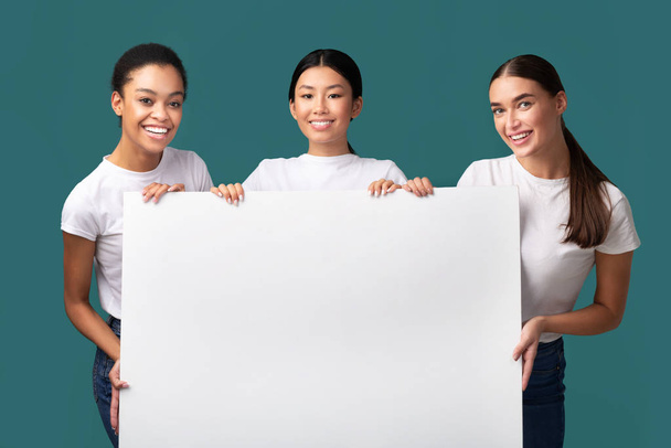 Drie vrouwen houden witte poster staande op turquoise achtergrond, Mockup - Foto, afbeelding