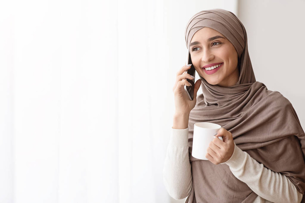 Happy Muslim Girl Talking On Cellphone And Drinking Coffee Near Window - Fotoğraf, Görsel