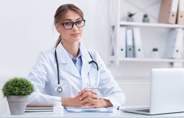 Female Doctor Looking At Camera Sitting At Workdesk In Office - Foto, Imagem