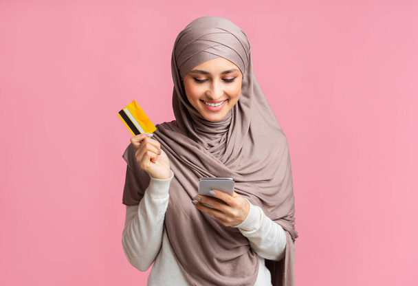 Mujer musulmana feliz en pañuelo para la cabeza con teléfono inteligente y la celebración de la tarjeta de crédito
 - Foto, imagen