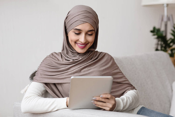 Femme musulmane souriante dans le hijab en utilisant une tablette numérique, se détendre à la maison
 - Photo, image