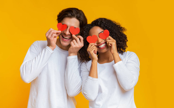 Romanttinen rotujenvälinen pari pitelee punaisia sydämiä silmien yli ja hymyilee
 - Valokuva, kuva