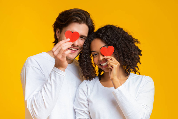 romantisches gemischtrassiges Paar, das Augen mit roten Papierherzen in Händen bedeckt - Foto, Bild