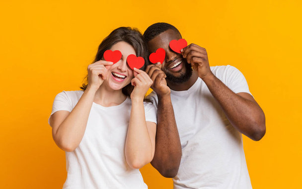 Romantic multiracial couple posing with red hearts over eyes and laughing - 写真・画像