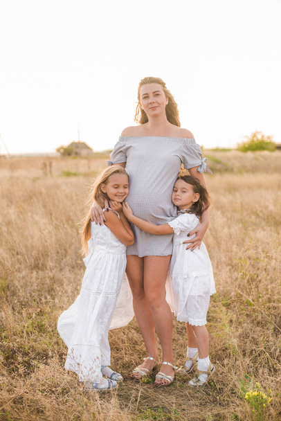 Beautiful young pregnant mother with blond hair and two daughters in white dresses in a field in the summer at sunset. Happy pregnancy. The large family. Family and Children - Fotó, kép