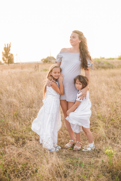 Beautiful young pregnant mother with blond hair and two daughters in white dresses in a field in the summer at sunset. Happy pregnancy. The large family. Family and Children - Foto, imagen