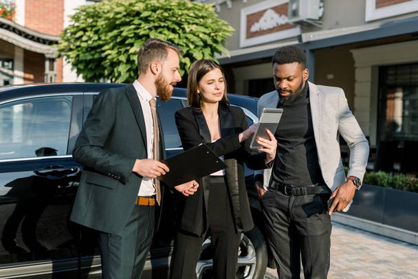 Two men, African and Caucasian, and one Caucasian woman stand in front of black car on a parking place on yard of dealer. Woman showing something on the digital tablet - Foto, Imagen