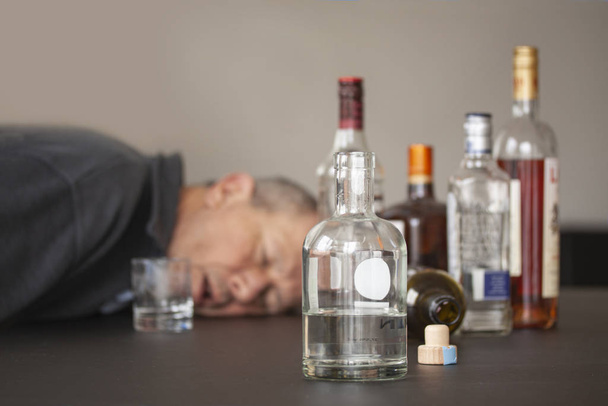 Μεθυσμένος άντρας που κοιμάται με το κεφάλι του ξαπλωμένο σε ένα τραπέζι και ανοιχτό στόμα και μπουκάλια αλκοόλ σκορπισμένα γύρω του. Ένα μπουκάλι γύρισε..  - Φωτογραφία, εικόνα