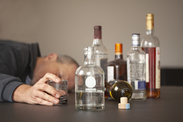 Drunken man sleeping with his head lying on a table and open mouth and alcohol bottles scattered around him. One bottle is flipped. His hand is holding on to an empty glass while in his sleep. - Photo, Image