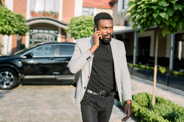 African american businessman in a suit speaking on smartphone, walking outdoors on the background of his black car and office buildings. - Zdjęcie, obraz