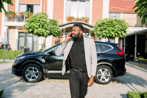 Serious rich African businessman in smart casual business suit talking over phone while walking on the yard and parking of modern business center. Black car crossover on the background - Foto, Bild