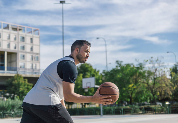 Joven y fuerte hombre juega baloncesto con su bola naranja con un hermoso cielo azul en el fondo
 - Foto, Imagen