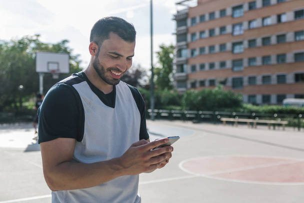 Attractive sportsman man looks at his mobile phone while taking a break in the middle of an urban court - Foto, immagini
