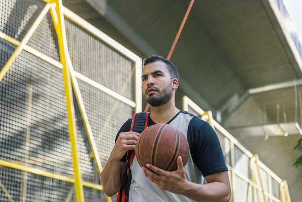 Detailní záběr pohledného a atraktivního mužského atleta latinského původu, nesoucího svůj basketbal při chůzi po ulici - Fotografie, Obrázek