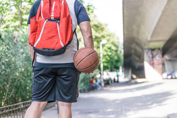 Back view of a male basketball player carrying his ball and a large red backpack on his back that walks through a park - Photo, Image
