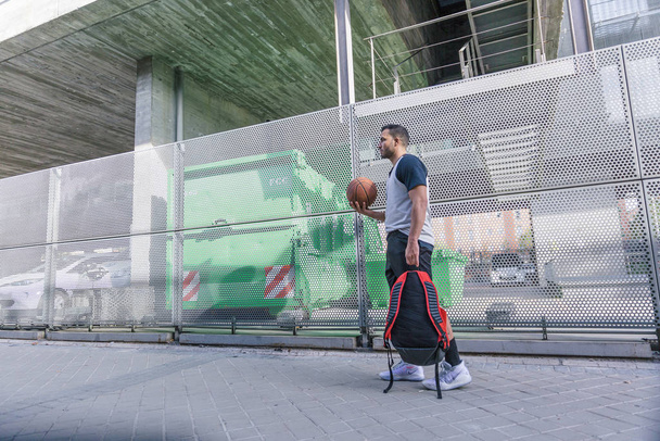Çekici bir basketbolcu kırmızı sırt çantası ve turuncu topuyla kentsel alanda yürüyor. - Fotoğraf, Görsel