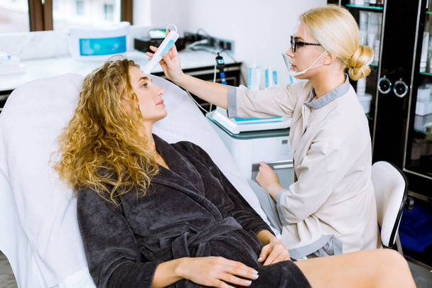 Pretty young blond curly woman during face skin diagnostic in the professional beauty salon with female doctor cosmetologist - Photo, Image