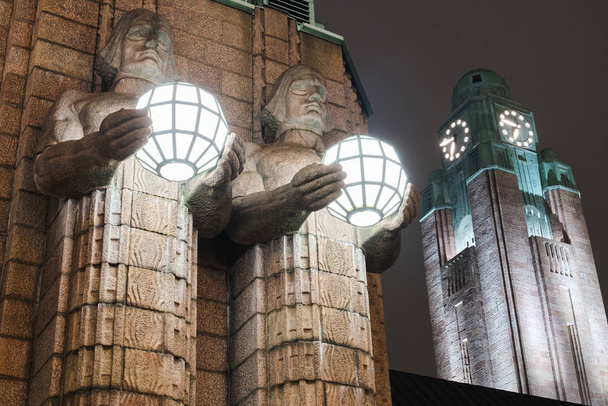 HELSINKI, FINLÂNDIA - NOVEMBRO 2019: Helsínquia, Finlândia. Vista noturna de dois pares de estátuas segurando as lâmpadas esféricas na entrada para a Estação Ferroviária Central de Helsinque. Iluminação noturna ou noturna
. - Foto, Imagem