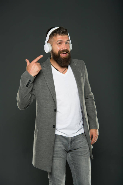 Look at my headset. Hipster point finger at headset grey background. Bearded man listen to music playing in stereo headset. Using wireless bluetooth headset for business and fun. Modern life - Foto, imagen