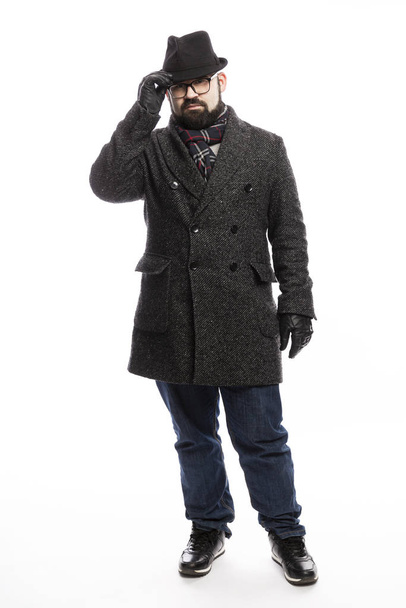 Elegant man with a beard in a coat and hat. He stands in full growth on a white background and holds on to his hat.  - Foto, Imagem