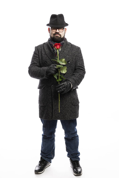 Elegantní muž s vousy, oblečený ve stylovém kabátě a klobouku s rudou růží v rukou. Plná výška. Izolované přes bílé pozadí.  - Fotografie, Obrázek