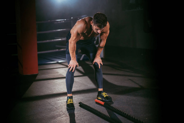 Харизматичный парень в спортивной одежде сгибает руки на коленях в спортзале. Фитнес-человек выглядит уставшим после интенсивной тренировки
 - Фото, изображение