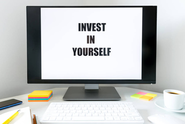 text displayed on the monitor, Invest in Yourself.  A desk with a keyboard, office accessories and a cup of coffee.  The concept of personal and professional development - Photo, Image