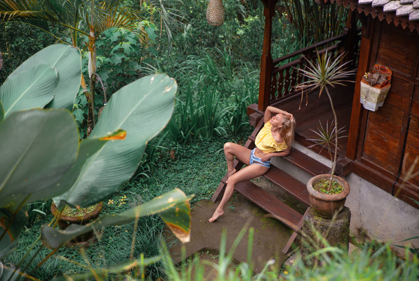 Γυναίκα με τζιν σορτς κάθεται στο τροπικό μπαλκόνι κοντά jungl - Φωτογραφία, εικόνα