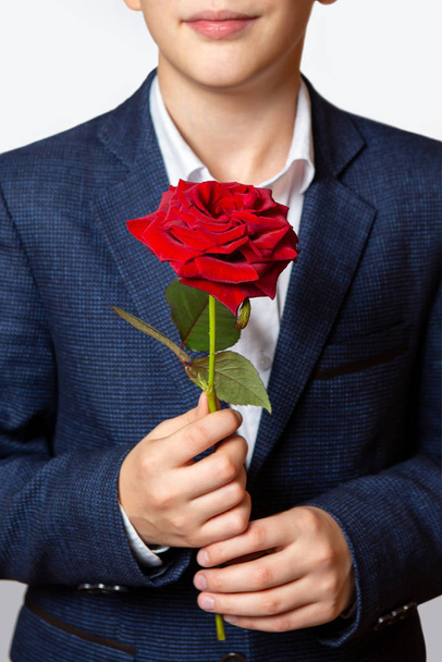 Red rose in the hands of a teenager. The boy is dressed in a blue suit and white shirt.  - Foto, afbeelding