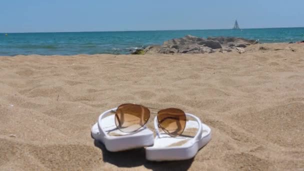 Damskie kapcie i okulary przeciwsłoneczne na plaży w pobliżu fal nad morzem - Materiał filmowy, wideo