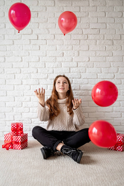 Linda joven sonriente sentada en la alfombra beige lanzando globos rojos al aire
 - Foto, imagen
