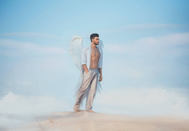 Handsome athletic man as an fallen angel walks sand desert blue sky cloud. Muscular sexy body naked torso beautiful body white shirt pants huge strong wings. fantasy design Creative costume clothes. - Foto, immagini