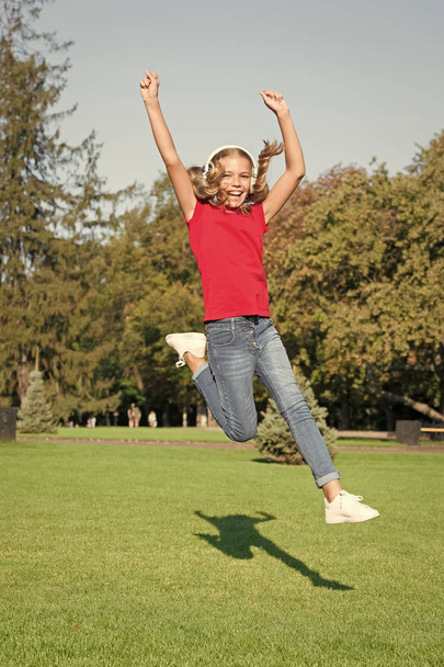 Energetic song that gets you moving. Energetic child jumping over green grass. Small girl enjoy energetic music in earphones. Active and energetic. Motion and freedom. Music and technology - Foto, Bild