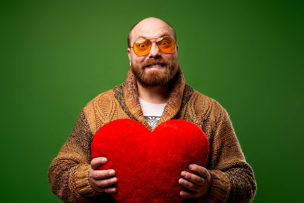 Man in pink glasses holds toy heart while standing on empty green background - Fotó, kép