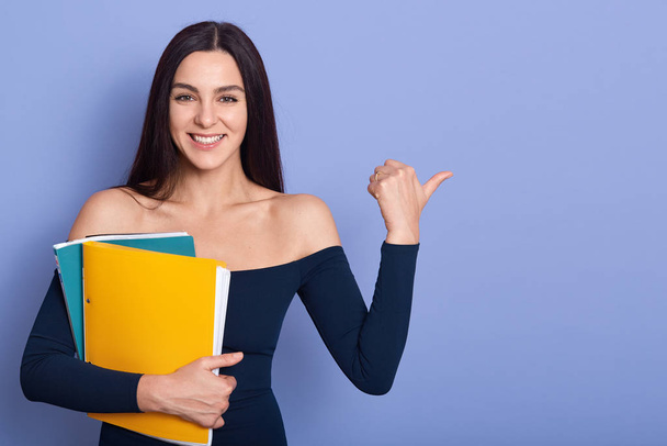 Caucasian businesswoman holding notepad and pointing aside with her thumb, elegance girl wearing dress with are shoulders, isoalted over blue background. Copynspace for advertisment or promotion. - Фото, изображение