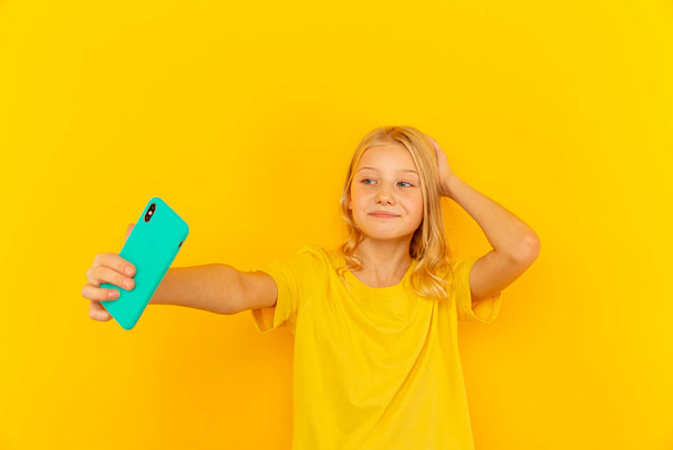 Χαμογελαστό μικρό κοριτσάκι δείχνει μπλε οθόνη του νέου δημοφιλούς κινητού τηλεφώνου σε ανοιχτό κίτρινο φόντο. - Φωτογραφία, εικόνα
