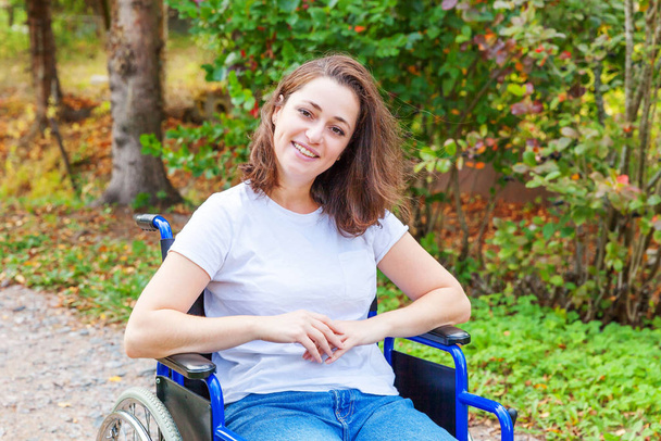 Молодая счастливая женщина-инвалид в инвалидной коляске на дороге в больничном парке ждет обслуживания пациентов. Парализованная девочка в инвалидном кресле для инвалидов на природе. Концепция реабилитации. - Фото, изображение