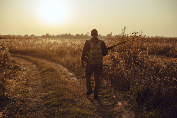 Fiatal vadász kockás ingben, katonai hátizsákkal és pisztollyal a kezében, amint a pályaúton sétál két mező között a naplementében - tökéletes alkalom kacsavadászatra. - Fotó, kép