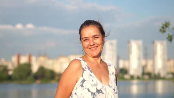 Portret van een gewone vrouw van middelbare leeftijd die glimlacht op de achtergrond van het meer. - Video