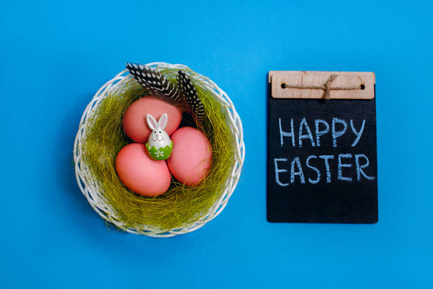 Пасхальный кролик лежит на розовых праздничных яйцах, на меловой тарелке Счастливая Пасхальная надпись, синий фон
 - Фото, изображение
