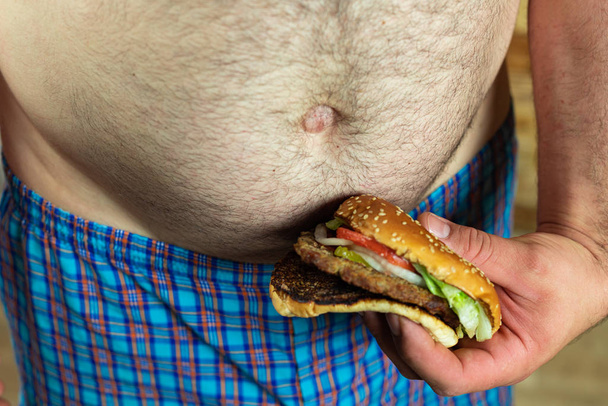 Man met heerlijke en sappige hamburger tegen zijn dikke buik. Anti-fastfood en tijd voor een dieetconcept. - Foto, afbeelding