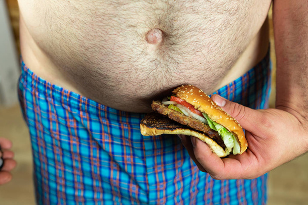 Man met heerlijke en sappige hamburger tegen zijn dikke buik. Anti-fastfood en tijd voor een dieetconcept. - Foto, afbeelding