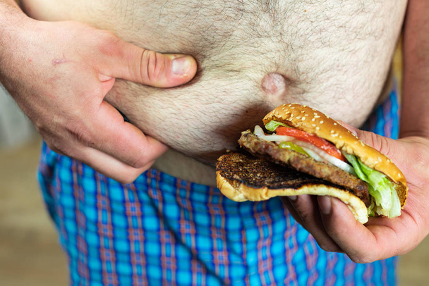 Hombre sosteniendo deliciosa y jugosa hamburguesa contra su vientre gordo. Anti comida rápida y tiempo para un concepto de dieta
. - Foto, Imagen