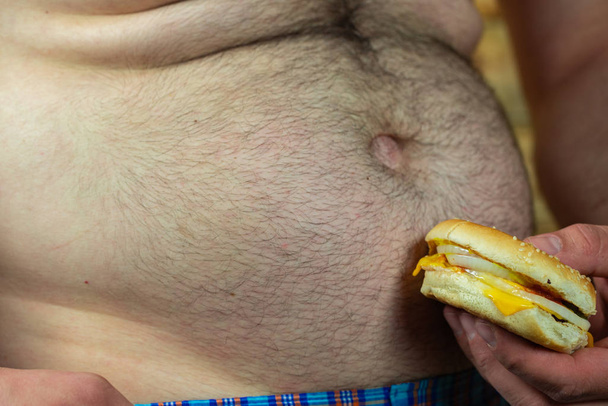 Homme tenant hamburger délicieux et juteux contre son gros ventre. Anti fast food et le temps pour un concept de régime
. - Photo, image